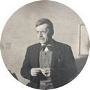 Une photographie aux tons sépia d'un homme portant un costume, un nœud papillon et un gilet, debout à l'intérieur avec une expression concentrée, tenant quelque chose de petit dans ses mains. Une photographie aux tons sépia d'un homme portant un costume, un nœud papillon et un gilet, debout à l'intérieur avec une expression concentrée, tenant quelque chose de petit dans ses mains.