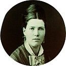 Portrait en noir et blanc d'une femme aux cheveux noirs coiffés en l'air, portant une tenue à col montant avec un nœud au cou et des boucles d'oreilles. L'image a un cadre ovale. Portrait en noir et blanc d'une femme aux cheveux noirs coiffés en l'air, portant une tenue à col montant avec un nœud au cou et des boucles d'oreilles. L'image a un cadre ovale.